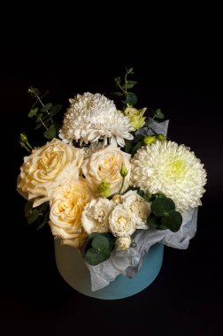 Yumuşak sarı güller ve beyaz kasımpatılardan oluşan güzel bir düzenleme, koyu siyah bir zemin üzerine kurulmuş, bir deniz mavisi kabın içinde sunuluyor..