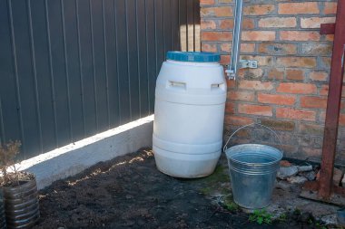 Beyaz bir su varili tuğla bir duvarın yanında duruyor. Yanında da dışarıda güneşin altında duran metal bir kova var..