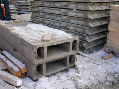 Kış inşaatı sırasında beton levhalar ve kirişler kısmen karla kaplı olarak dışarıda yığılır. Arka planda bir kişi duruyor..