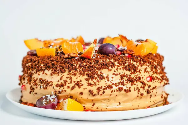 Taze meyve, fındık ve çikolata parçacıklarıyla süslenmiş lezzetli bir pasta. Pasta, beyaz bir tabakta, gözlere hitap eden beyaz bir zemin üzerinde sunuluyor..