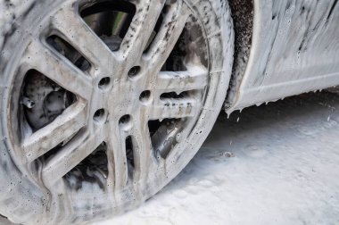 Bir araba tekerleği, profesyonel bir araba yıkama sırasında kalın, beyaz sabunlarla kaplıdır, su damlacıkları görünür..