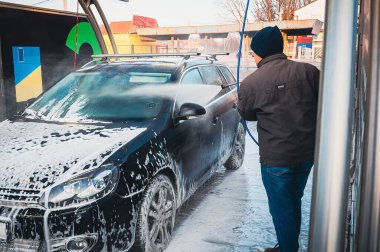 Oto yıkamada siyah bir arabayı özenle yıkayan bir adam, yüksek basınçlı su hortumuyla iyice duruluyor, köpükler her yerde..