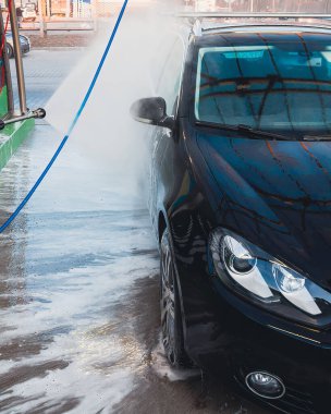 Parlak siyah bir araba bir oto yıkama istasyonunda yüksek basınçlı su ve köpükle yıkanmaktadır. Baloncuklar ve spreyler temiz görünümü arttırır..