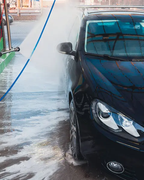 Parlak siyah bir araba bir oto yıkama istasyonunda yüksek basınçlı su ve köpükle yıkanmaktadır. Baloncuklar ve spreyler temiz görünümü arttırır..