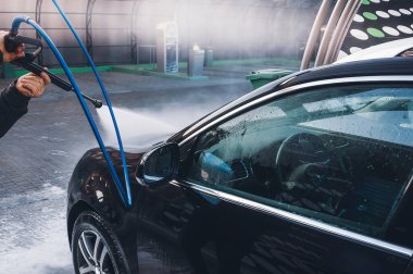 Parlak bir günde oto yıkama istasyonunda yüksek basınçlı su püskürtücüsüyle siyah bir arabayı yıkayan biri..