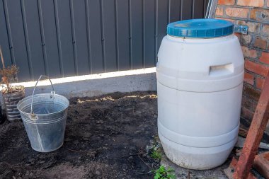 Tuğla bir duvarın yanında beyaz bir su varili ve bahçe düzeninde metal bir kova duruyor. Kova yerde duruyor..