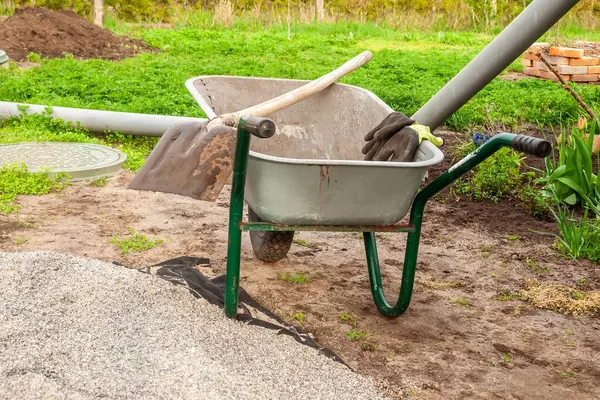 Bir el arabası eldiven ve aletlerle oturur, bahçe işleri için hazırdır. Onun yanında da çakıl ve yemyeşil otlar vardır..