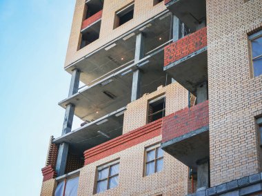 Yakın çekimde, mavi gökyüzü zeminine karşı kırmızı ve bej tuğlalı inşaat halindeki bir bina görülüyor.