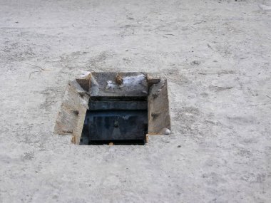 Beton bir yüzeyde açık bir rögar kapağı var. Altındaki karanlık açıklığı ortaya çıkarıyor. Sahne kentsel altyapı öneriyor.