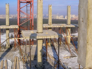 Beton sütunları ve vinci olan bir inşaat sahası, arka planda şehir silüeti olan, gün ışığında.