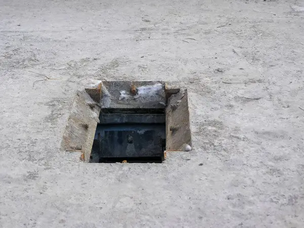 Beton bir yüzeyde açık bir rögar kapağı var. Altındaki karanlık açıklığı ortaya çıkarıyor. Sahne kentsel altyapı öneriyor.