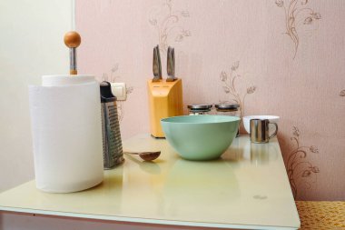 Mutfak sahnesinde rende, bıçak ve kase gibi çeşitli aletler var. Çiçekli duvar kâğıdı arkaplanı olan bir masaya dizilmiş..