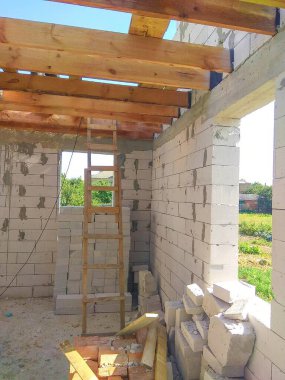 Ahşap kirişleri ve merdiveni olan bir inşaat alanı. Bina yapım aşamasında, blok duvarlar ve görünür bir çatı yapısı var, gün ışığında..