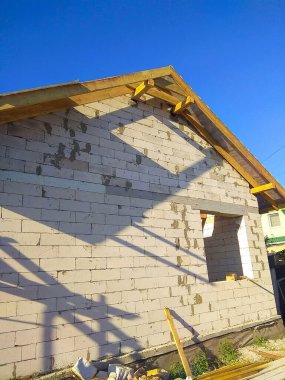 Açıkta tuğla duvarlar, ahşap kirişler ve açık mavi gökyüzü olan inşaat halindeki bir ev inşaat sürecini gözler önüne seriyor..