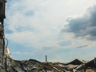 Hasarlı bir binanın kalıntıları parçalı bulutlu bir gökyüzünün altında uzun bir baca ile görülür..