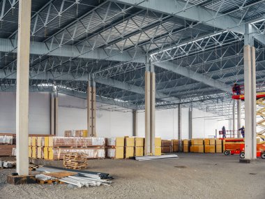 Metal çatı yapısı, beton sütunlar ve malzeme yığınlarının yer aldığı büyük bir sanayi deposunun içinde..