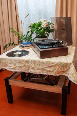 Klasik bir turntable çiçekli masa örtüsüyle kaplı bir masanın üzerinde oturur. Bitkiler ve dekorla nostaljik ve samimi bir atmosfer yaratır..