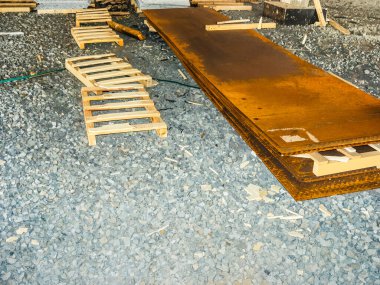 İnşaat alanındaki ahşap paletlerin üzerine yığılmış paslı metal levhalar. Yer çakıllarla kaplı..