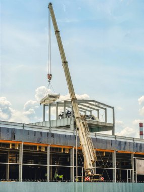 İnşaat vinci, bulutlu bir gökyüzüne karşı inşaat halindeki bir binaya büyük metal bir panel kaldırıyor..