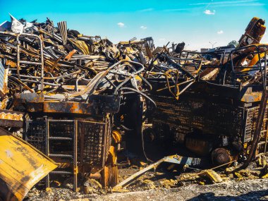 Bir aracın enkazının detaylı bir görüntüsü, muhtemelen bir inşaat ya da endüstriyel makine, yangından sonra, mavi gökyüzü altında kömürleşmiş metal ve enkaz ile.