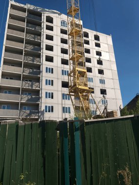 İnşaat halindeki yüksek bir bina, bitmemiş yapının üzerinde yükselen sarı bir vinç, açık mavi bir gökyüzüne karşı..