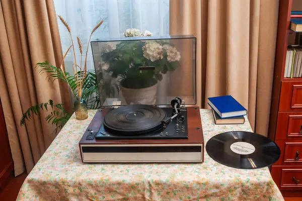 Eski bir plak çalar çiçeklerin olduğu bir masa örtüsünün üzerinde oturur, yanında plaklar ve kitaplar vardır. Beyaz çiçekli bir bitki sahnenin estetiğine katkı sağlar..