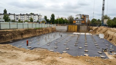 Temel direkleri olan bir inşaat alanı. Arkaplanda bulutlu bir gökyüzünün altında bir vinç ve binalar görülebilir..