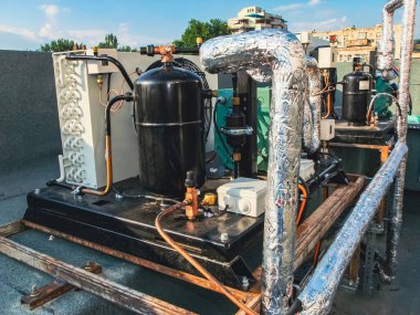 Endüstriyel bir çatı havalandırma ünitesi, sergilenen borular, bir kompresör ve diğer mekanik bileşenler, parlak, güneşli bir zemin üzerine kurulmuş..