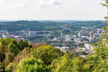 Binalar, ağaçlar ve parlak gökyüzü ile yemyeşil tepeler arasında yuva yapmış bir şehrin hava manzarası..