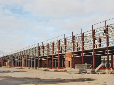 A long industrial building under construction, featuring steel framework and brick walls, with workers visible on the structure. The sky is overcast.
