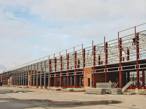 A long industrial building under construction, featuring steel framework and brick walls, with workers visible on the structure. The sky is overcast.