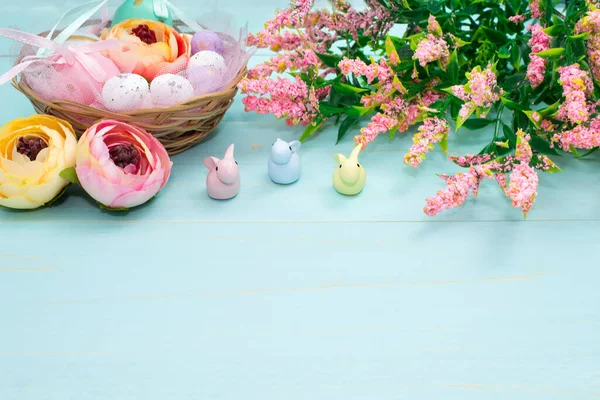 Easter composition of pink spring flowers, colorful roses, cute bunnies and decorative eggs in a nest. Content for Easter holiday on blue wooden background. Flat lay, side view, close up, copy space