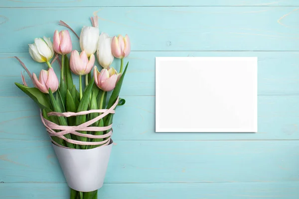 Composition of pink and white tulips, ribbons, blank card on blue wooden background. Tulips spring bouquet. Content for Birthday, Valentines Day, Womens day. Flat lay, top view, close up, copy space.