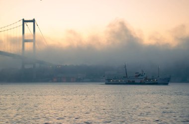 15 Temmuz Şehitler Köprüsü 'nün sisli bulutlarda martı ve gündoğumu manzarası. Şehir hatları feribotu Sis Boğazı 'nda ve Boğaz Köprüsü' nün altında hareket ediyor..