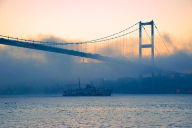 15 Temmuz Şehitler Köprüsü 'nün sisli bulutlarda martı ve gündoğumu manzarası. Şehir hatları feribotu Sis Boğazı 'nda ve Boğaz Köprüsü' nün altında hareket ediyor..