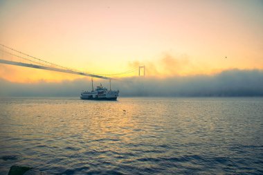 15 Temmuz Şehitler Köprüsü 'nün sisli bulutlarda martı ve gündoğumu manzarası. Şehir hatları feribotu Sis Boğazı 'nda ve Boğaz Köprüsü' nün altında hareket ediyor..