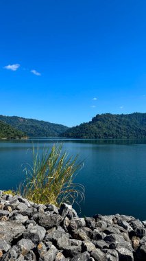 Tayland, Nakhon Nayok 'daki Khun Dan Prakarnchon Barajı' ndan rezervuarın panoramik manzarası ve çevresindeki yemyeşil manzara. Sakin su, yeşil tepeler ve ormanlarla çevrili temiz gökyüzünü yansıtır.