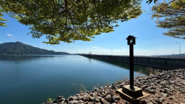 Tayland, Nakhon Nayok 'daki Khun Dan Prakarnchon Barajı' ndan rezervuarın panoramik manzarası ve çevresindeki yemyeşil manzara. Sakin su, yeşil tepeler ve ormanlarla çevrili temiz gökyüzünü yansıtır.