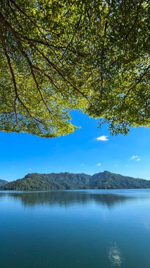 Tayland, Nakhon Nayok 'daki Khun Dan Prakarnchon Barajı' ndan rezervuarın panoramik manzarası ve çevresindeki yemyeşil manzara. Sakin su, yeşil tepeler ve ormanlarla çevrili temiz gökyüzünü yansıtır.