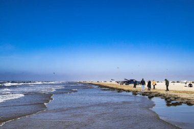 an adventurous beach with parked cars right by the sea, athletes, walkers and flying kites on the island of Rm in Denmark clipart