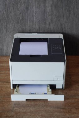 Ahşap zeminde duran siyah-beyaz lazer yazıcı. Yazıcının üstündeki ana kağıt çıkış tepsisinde bir yığın beyaz kağıt görünür ve alt kağıt girdi tepsisi çıkarılır.                                