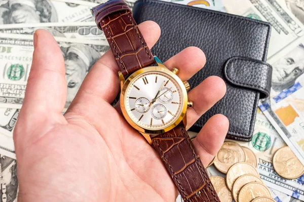 Hand holding wrist watch on background of dollars and wallet.