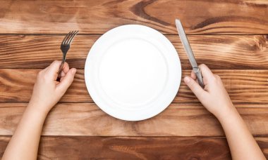 Aç çocuk yemek bekliyor. Çocuğun elleri çatal ve bıçağı boş tabakla masanın üzerinde tutuyor. Üst görünüm.