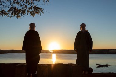 Two arab young man backward wearing djellaba and turban looking to the sunset in front of a lake in clear sky
