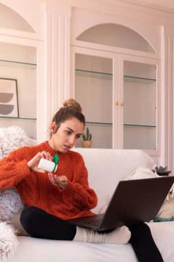 Evdeki karantina sırasında laptopta doktorla çevrimiçi sohbet ederken koltukta oturup plastik konteynırdan haplar alan sıcak kanlı genç bir kadın.