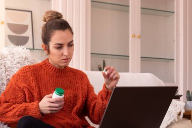 Bilgisayardaki randevuda kapsülü doktorlara gösterirken koltukta elinde plastik haplarla oturan sıcak süveter giymiş endişeli genç bir kadın.