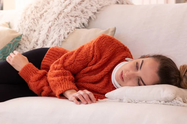 Tired young woman in orange warm clothes with orthopedic collar lying down on couch to rest after cervical injury in living room at home