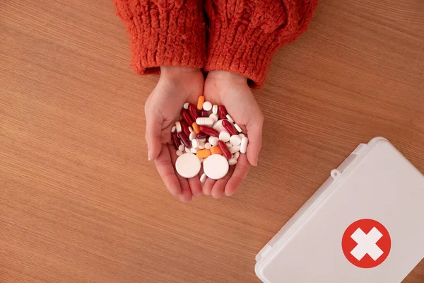 Top view of crop unrecognizable female in knitted sweater showing handful of colorful pills near first aid kit