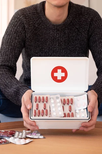Crop unrecognizable man in casual outfit sitting in light room and showing first aid kit with cross and different pills