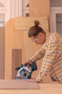 Gözlüklü bir erkek marangozun atölyede mobilya yaparken ahşap tahtayı dairesel testereyle keserken yan görünüşü